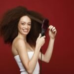 Against a vibrant red backdrop, a woman with a dazzling smile effortlessly tames her voluminous curls using a TYMO hair brush, showcasing the fun and ease of transforming her look.