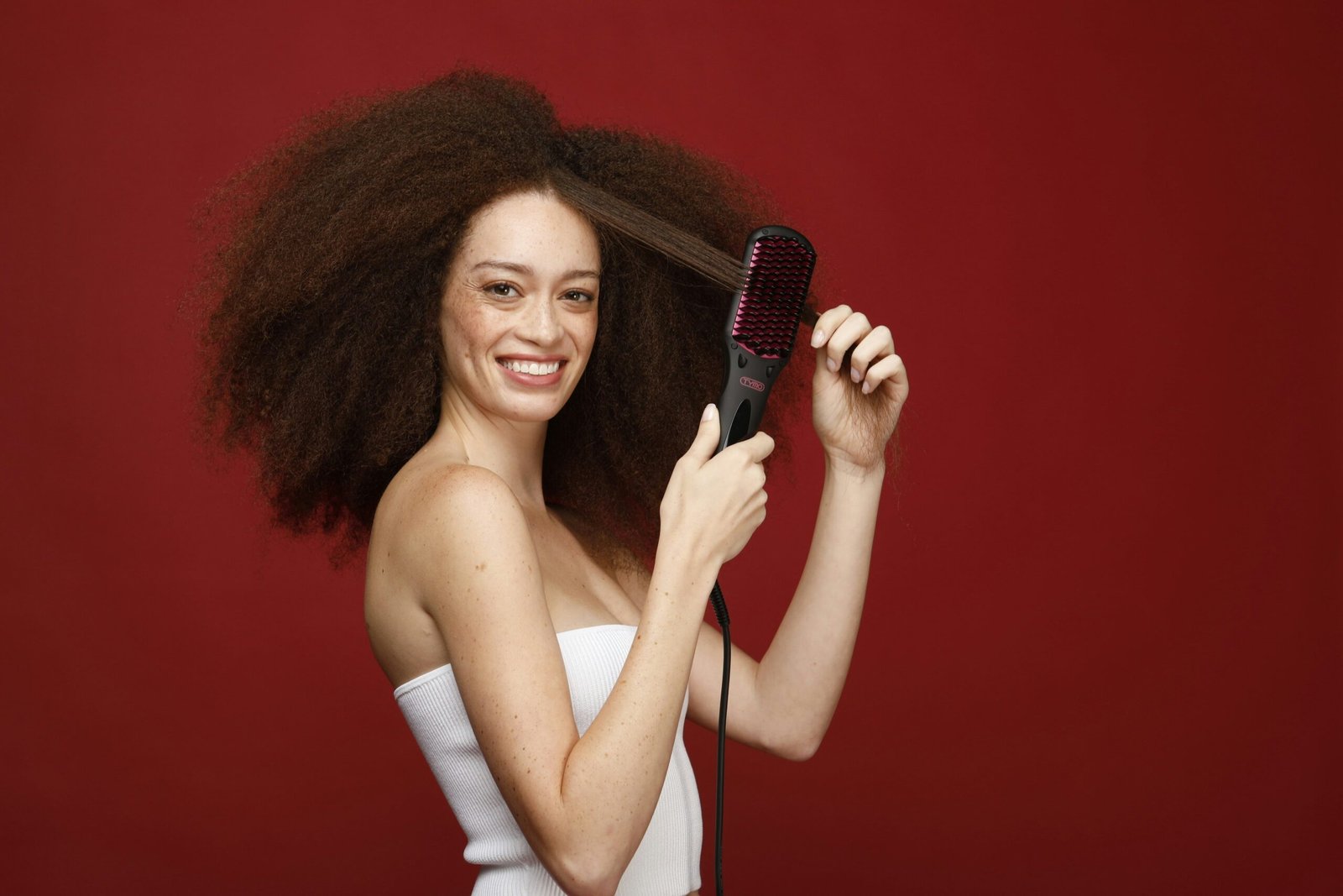 Against a vibrant red backdrop, a woman with a dazzling smile effortlessly tames her voluminous curls using a TYMO hair brush, showcasing the fun and ease of transforming her look.