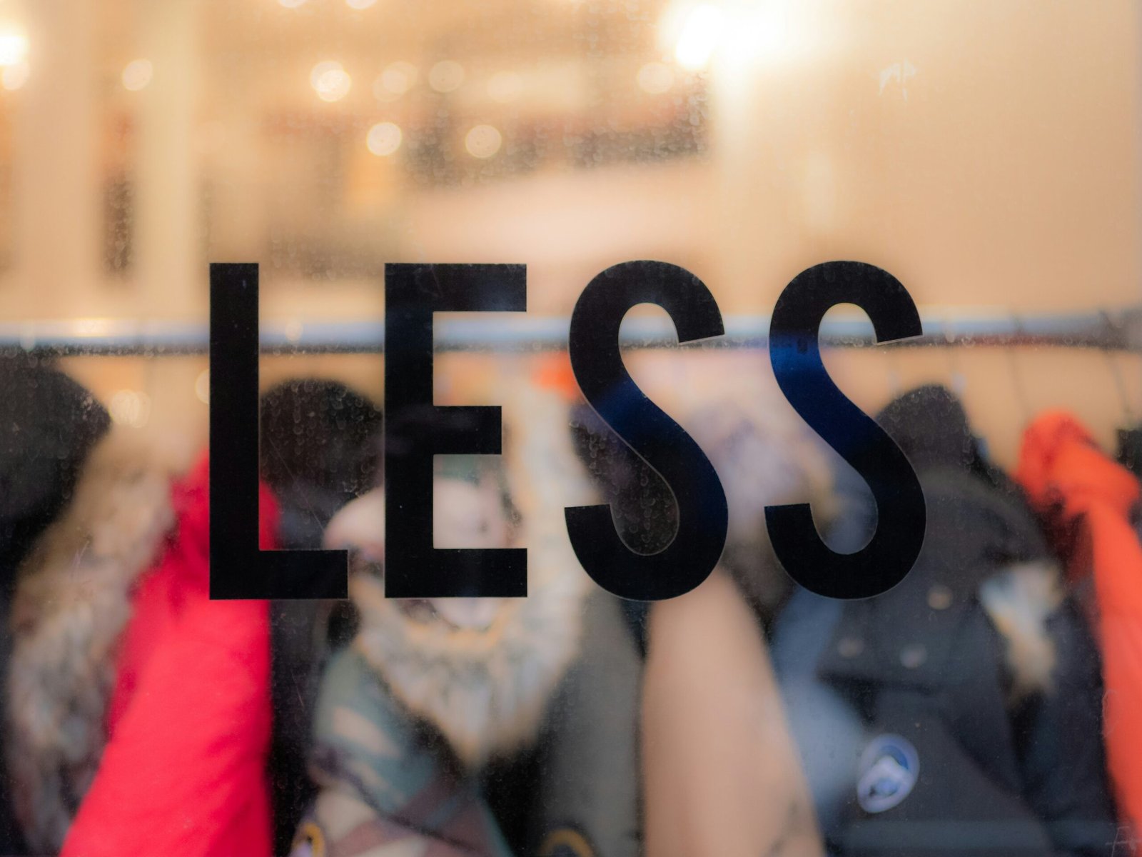 The word “less” on a window, in focus with clothes behind it. Commentary on consumerism and waste in the fashion industry.