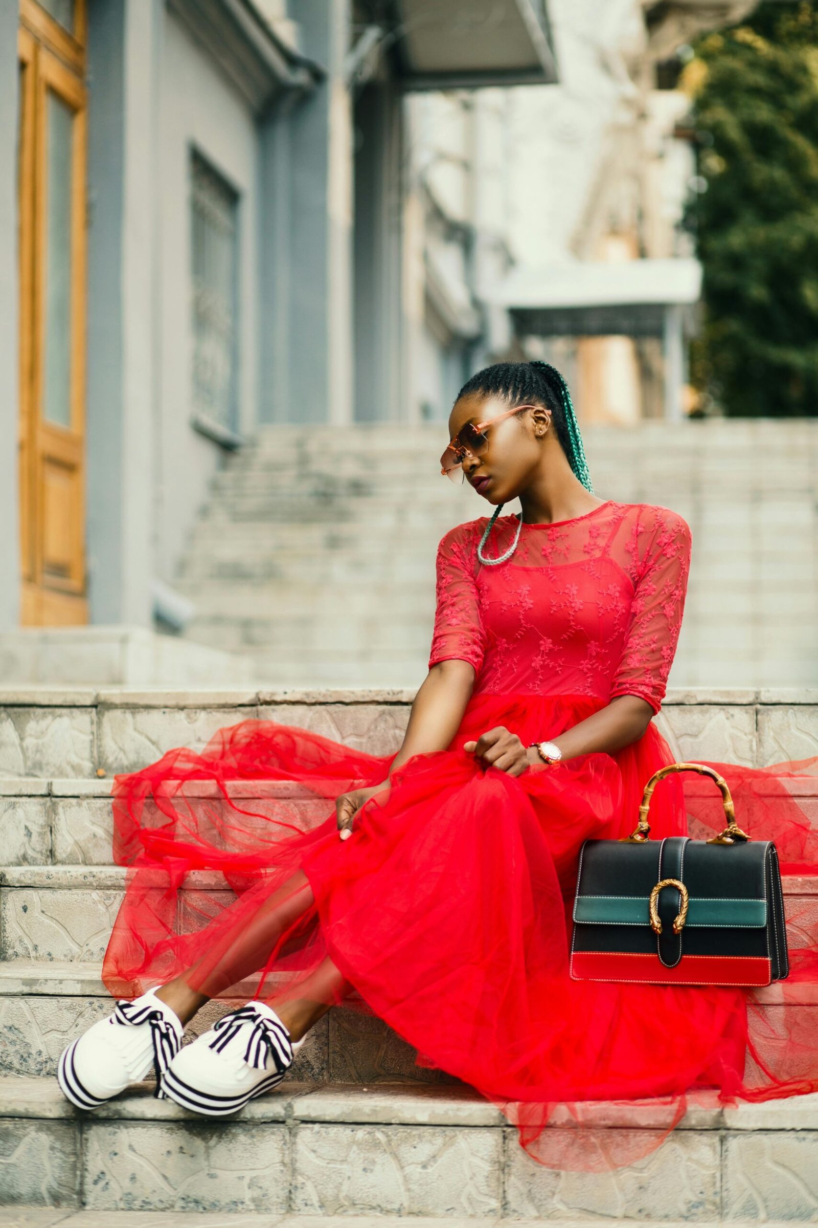 Woman wearing red long sleeved dress near black leather bag sitting on concrete stairs posing for photoshoot